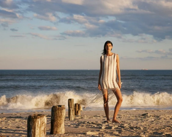 Model Rossana Latallada posing on a sandy beach wearing a striped sleeveless tunic dress with fringe details by breaking ocean waves.