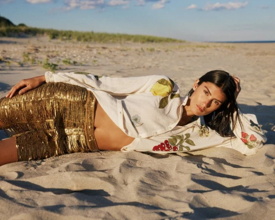 Model Rossana Latallada reclines on a sandy beach wearing metallic gold shorts and an embroidered cream fruit shirt.