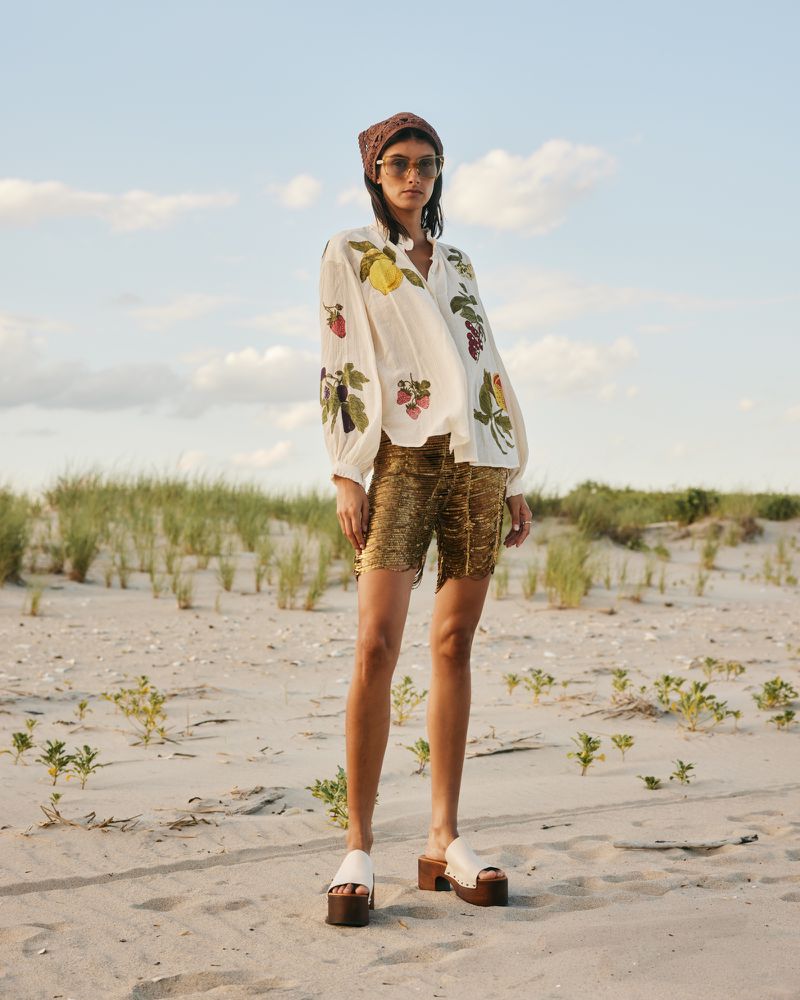 Model Rossana Latallada posing on sandy beach dunes wearing a fruit-embroidered blouse, gold shorts, and platform clogs.