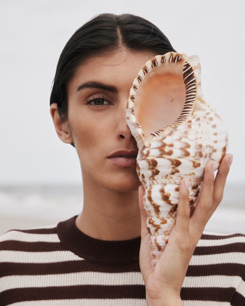 Model holding a large patterned seashell over half her face while wearing a brown and cream striped knit sweater on a beach.