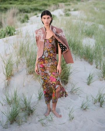 Model Rossana Latallada standing barefoot on grassy sand dunes in a floral dress and brown embroidered jacket.