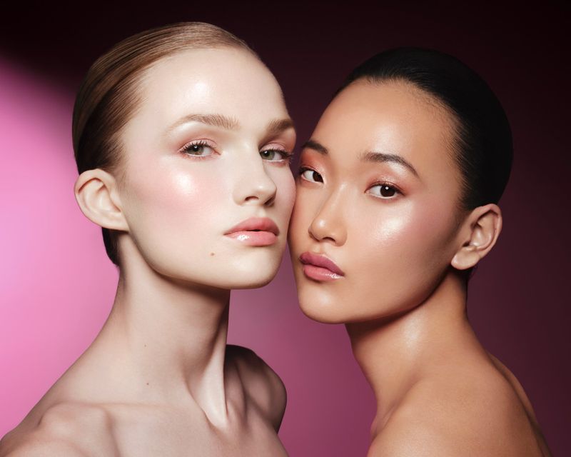Two women, one fair-skinned and one Asian, pose cheek-to-cheek with glossy makeup against a gradient pink backdrop in a beauty shot.