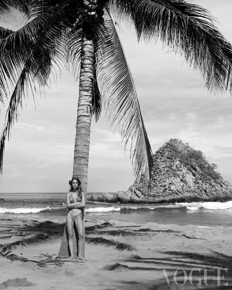 Maria Klaumann stands beside a tall palm tree in a bikini on a sandy beach in a black and white editorial shot.