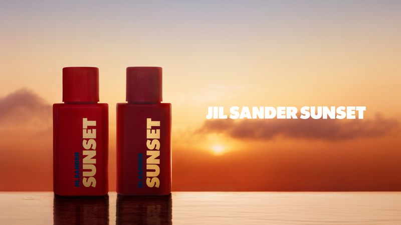 Two red Jil Sander Sunset perfume bottles are set against a vibrant orange and yellow sunset background. Minimalist product shot.