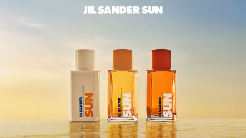 Three JIL SANDER SUN fragrance bottles in white, orange, and red tones on a reflective surface against a blue sky background.