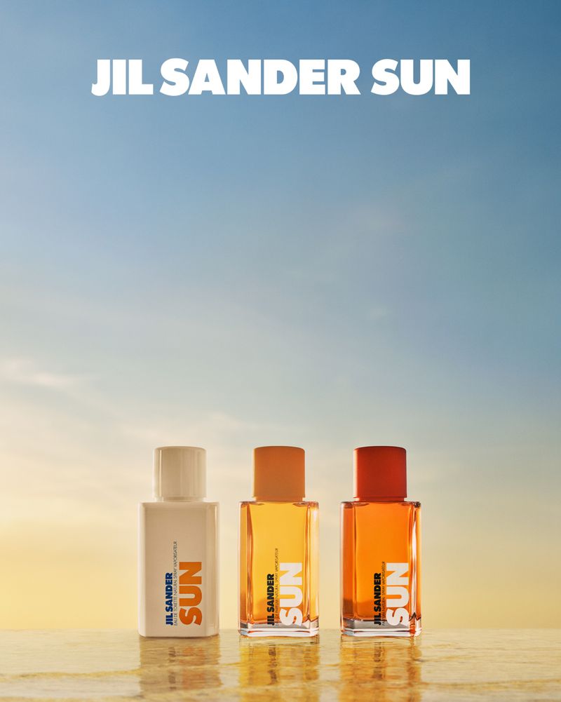 Three JIL SANDER SUN fragrance bottles stand on a reflective surface against a gradient sky, showcasing a minimal and summery aesthetic.