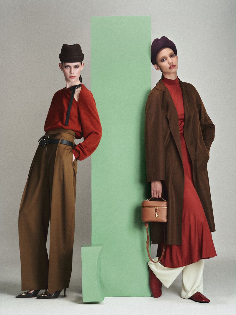 Cora Emmanuel and a model pose in tailored brown trousers, red knitwear, and hats by a green column for Elle Germany.