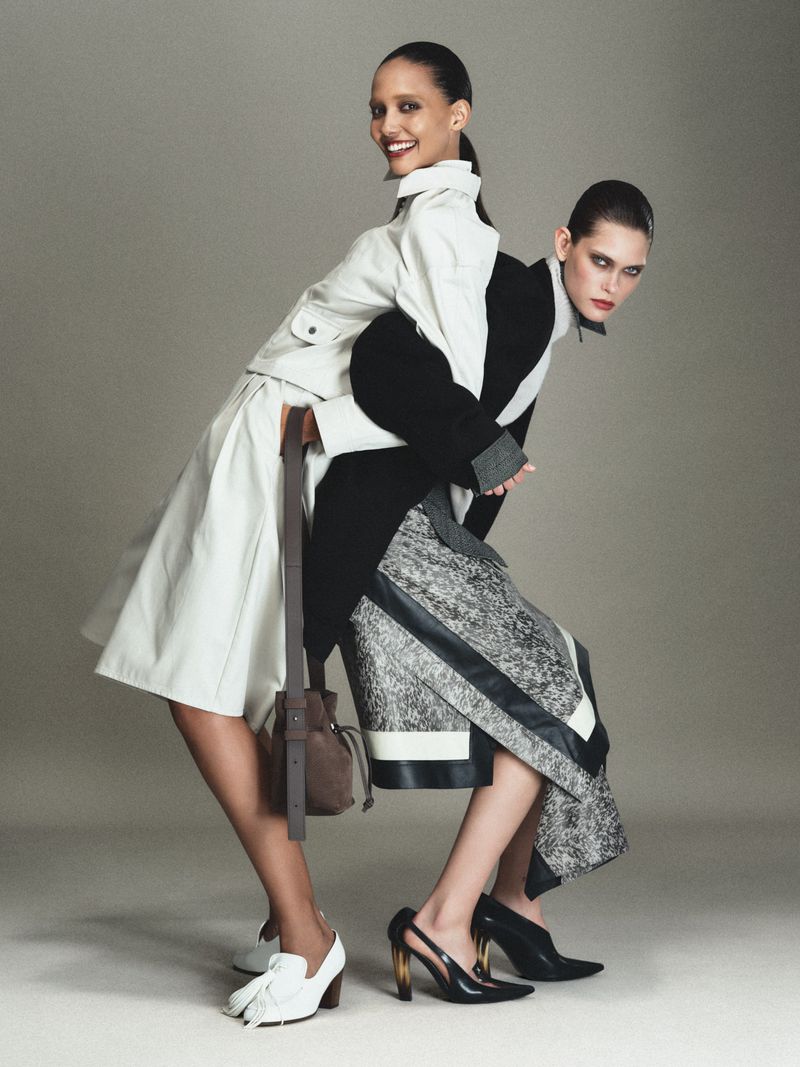 Two fashion models including Cora Emmanuel posing back to back in tailored monochrome outfits against a neutral studio backdrop.