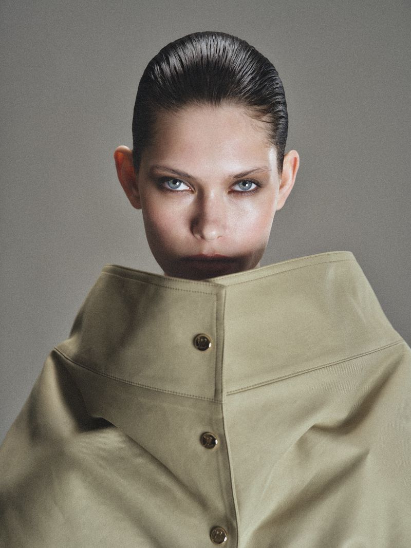 Model with slicked-back hair wearing a structured beige high-collar coat with round buttons against a neutral grey studio backdrop.