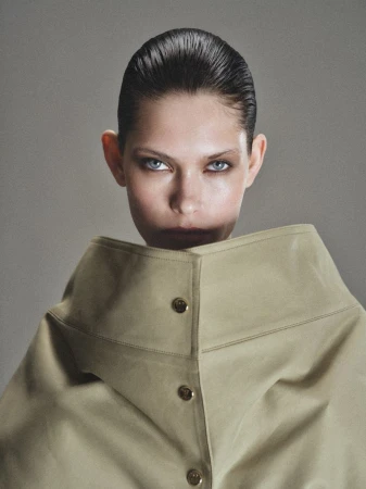 Model with slicked-back hair wearing a structured beige high-collar coat with round buttons against a neutral grey studio backdrop.