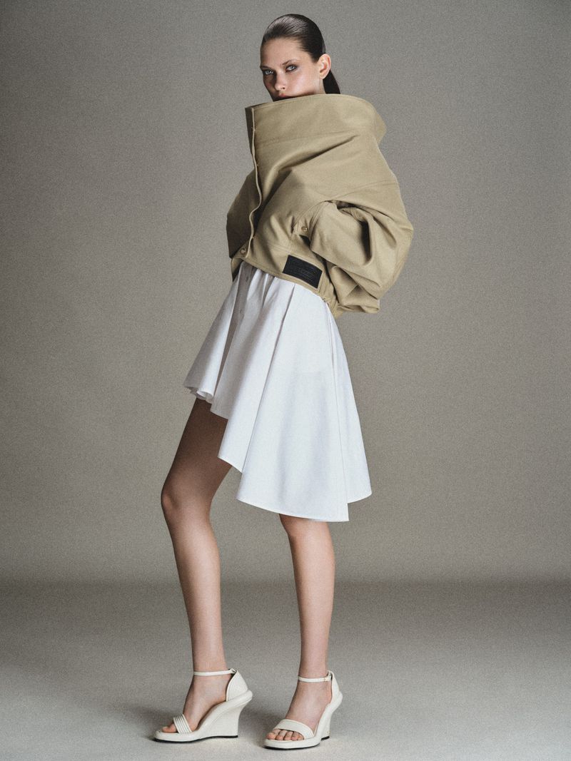 Model wearing an oversized beige collared jacket and a white pleated skirt against a grey backdrop