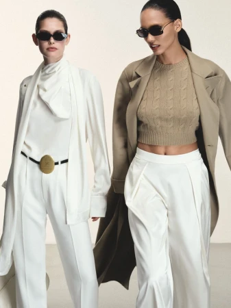 Two fashion models including Cora Emmanuel walking in tailored white suits and camel coats with dark sunglasses against an off-white backdrop.
