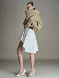 Model wearing an oversized beige collared jacket and a white pleated skirt against a grey backdrop
