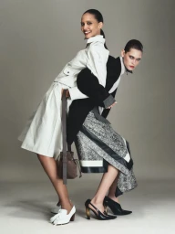 Two fashion models including Cora Emmanuel posing back to back in tailored monochrome outfits against a neutral studio backdrop.