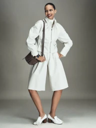 Model Cora Emmanuel posing in a white jacket, matching culottes, and white heeled loafers against a grey studio backdrop.