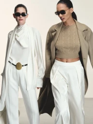 Two fashion models including Cora Emmanuel walking in tailored white suits and camel coats with dark sunglasses against an off-white backdrop.