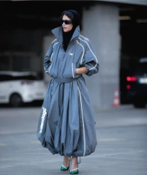 Model wearing an oversized grey Balenciaga track dress with a high collar, black hood, and emerald green heels in a carpark.