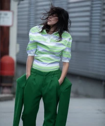 A woman in a green-and-white striped polo shirt and tailored green trousers standing outdoors with windblown dark hair.