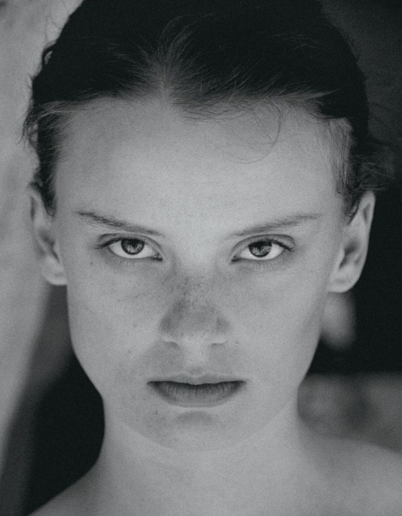 Black and white portrait of a woman with tied back hair and freckles staring directly at the camera in a close-up headshot.