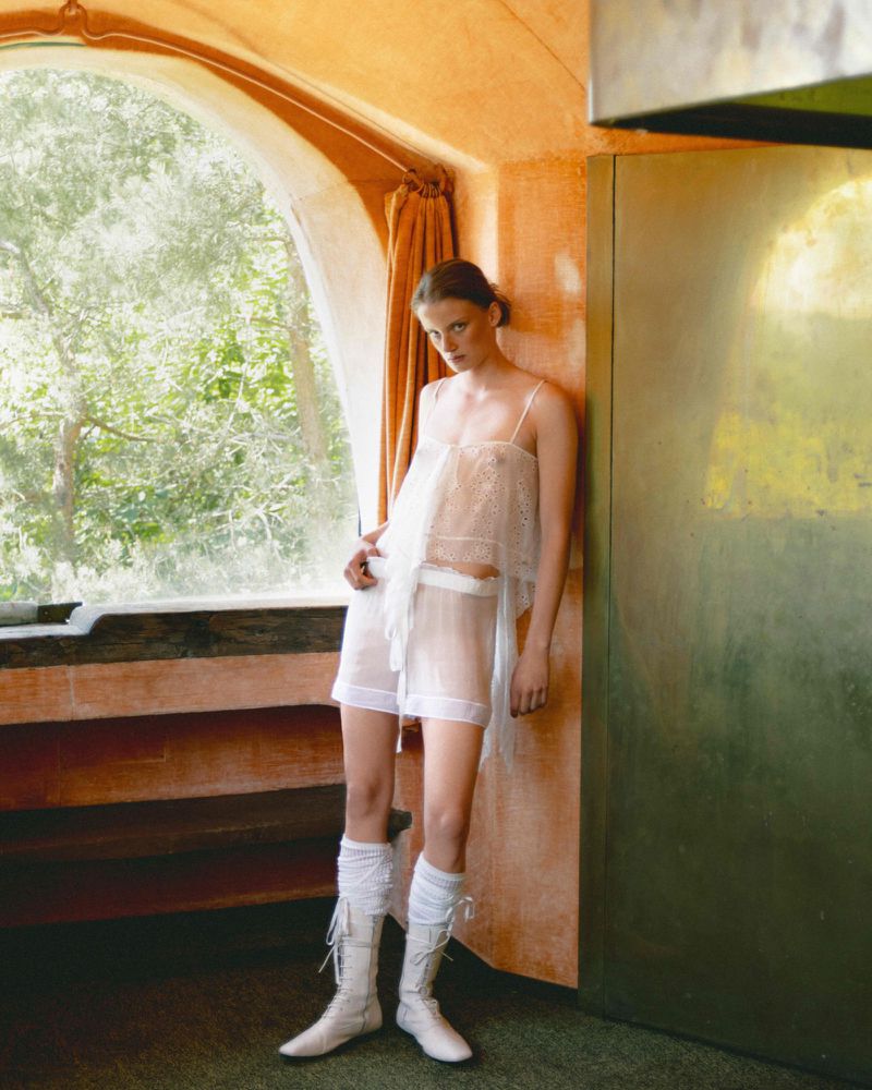 Indoor shot of a young adult woman in a sheer top and short skirt, leaning against a wall with a large window and wearing knee-high boots.
