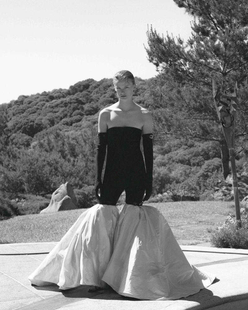 Model posing outdoors in a black strapless top, black gloves, and a white skirt, standing on a concrete patio with a wooded hillside.