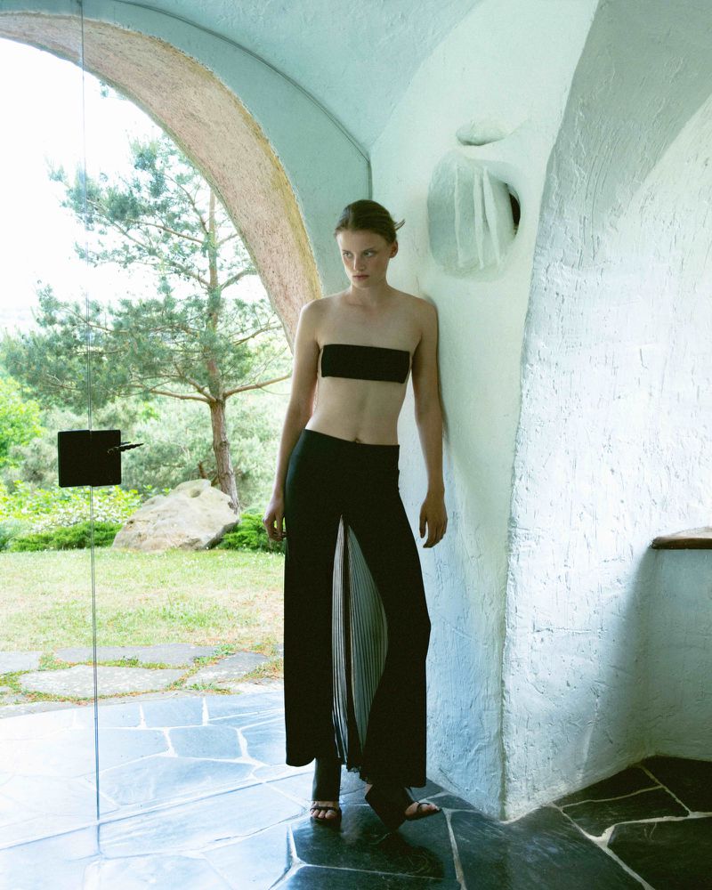 A slim adult woman in a black bandeau top and split-front trousers poses inside a light-filled, whitewashed, arched building exterior.
