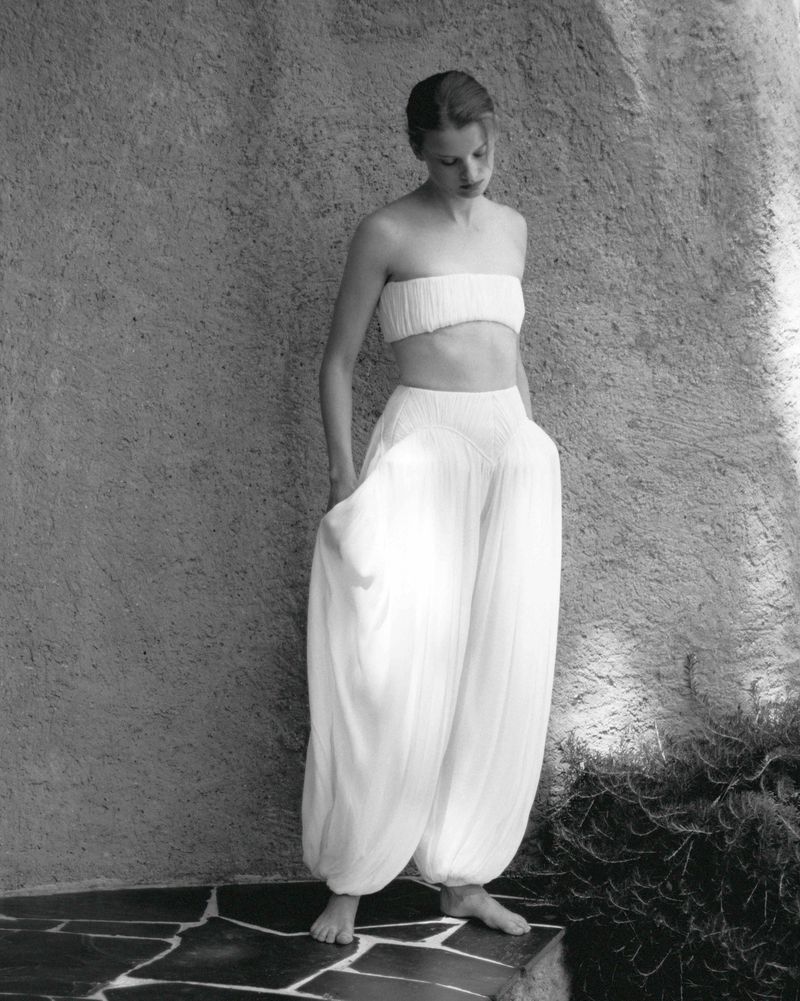Young woman poses in a white two-piece outfit with billowing harem pants against a textured stone wall in an outdoor setting.
