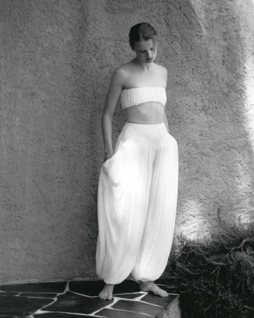 Young woman poses in a white two-piece outfit with billowing harem pants against a textured stone wall in an outdoor setting.