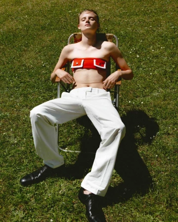 A fair-skinned young adult is reclining on a lawn chair, wearing a red top, white pants, and black shoes in a sunny outdoor setting.