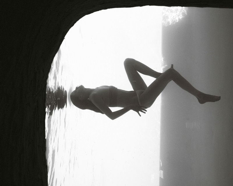 An adult woman in a bikini floats underwater in a pool of blue light, beneath a dark cavern or grotto, creating a serene, surreal image.