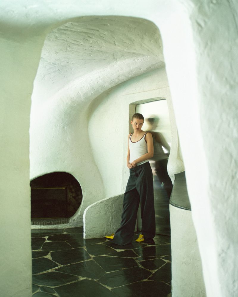 Indoor shot of a young woman wearing a white tank top and dark, wide-leg pants, standing in a room with a black-tiled floor.