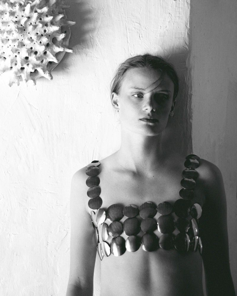 Monochrome portrait of a woman with shoulder-length hair wearing a disc-shaped metal top, posing against a textured wall.