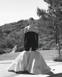 Model standing outdoors wearing a strapless black jumpsuit with dramatic voluminous trousers against a hilly landscape