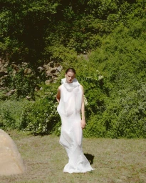 Model standing outdoors in a white sculptural dress against a dense green foliage and grass background.