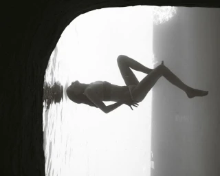 Monochrome silhouette of a woman lying down inside a dark cave opening looking out towards bright water