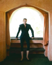 Model standing framed by an arched window wearing a dark tailored suit and cropped trousers