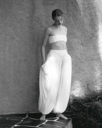 Model standing outdoors wearing white strapless bandeau top and voluminous high-waisted trousers against a textured stone wall