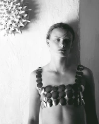 Black and white portrait of a woman wearing a metallic circular disc bralette against a textured plaster wall with a spiky wall sculpture.