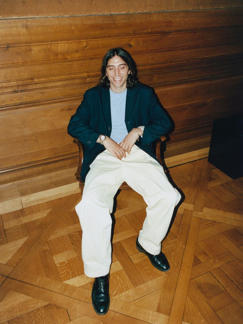 A man in a dark navy blazer and wide cream trousers smiling while seated in a wooden armchair on parquet flooring.