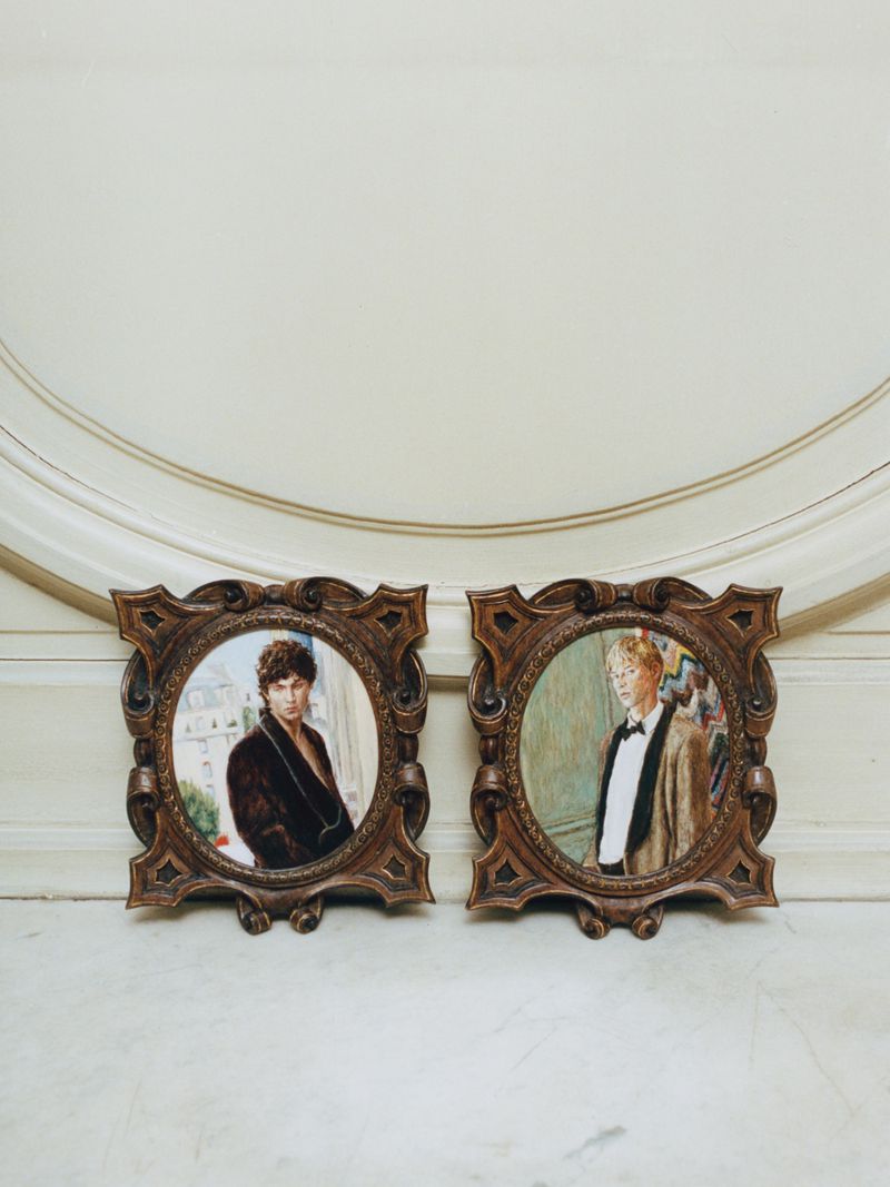 Two ornate carved miniature portrait frames featuring painted portraits of men in tailoring resting against an off-white wall.