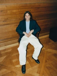 A man in a dark navy blazer and wide cream trousers smiling while seated in a wooden armchair on parquet flooring.