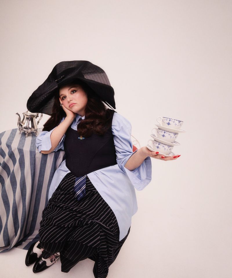 Woman poses in an eclectic outfit with a striped skirt, blue button-down shirt, and oversized hat, holding a stack of teacups.