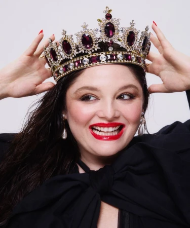 Close-up portrait of a smiling adult female wearing a jeweled crown and black dress against a white background in a photo studio.