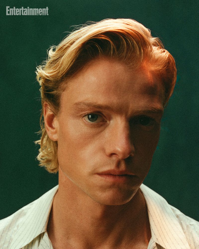 Close-up portrait of actor Tom Rhys Harries in a white collared shirt against a dark green background.