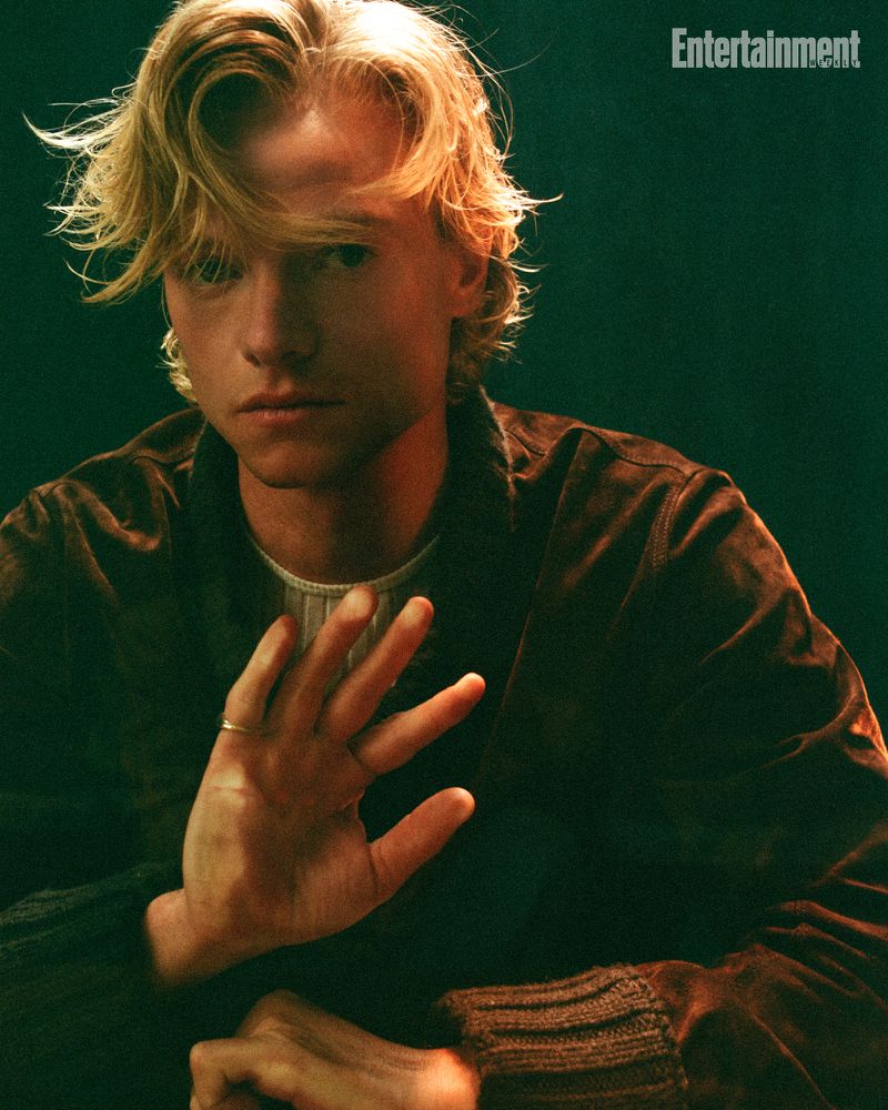Actor Tom Rhys Harries wearing a brown suede jacket raising his hand toward the camera in a moody portrait for Entertainment Weekly.