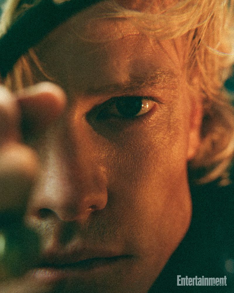 Extreme close up portrait of actor Tom Rhys Harries with blonde hair looking directly forward with hand blurred in foreground.