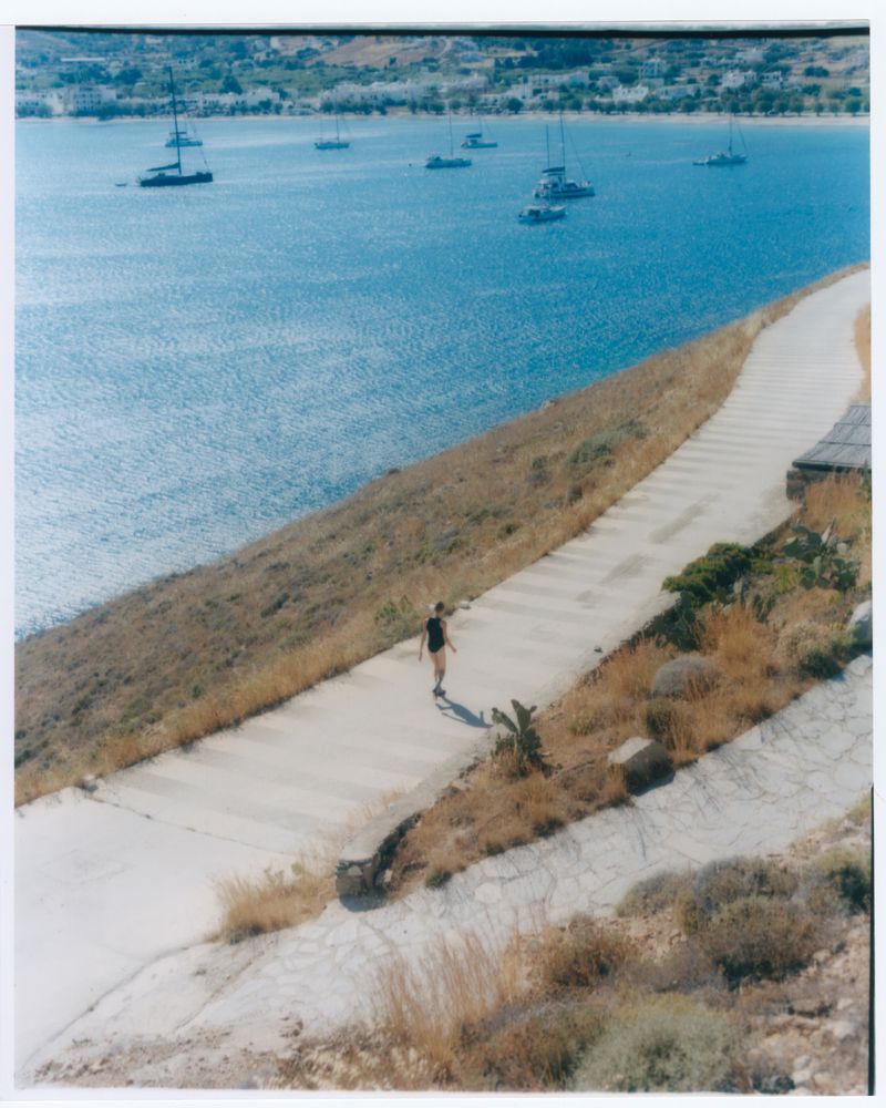 Person walking along a concrete path next to the ocean, with several sailboats on the water and land visible in the distance.