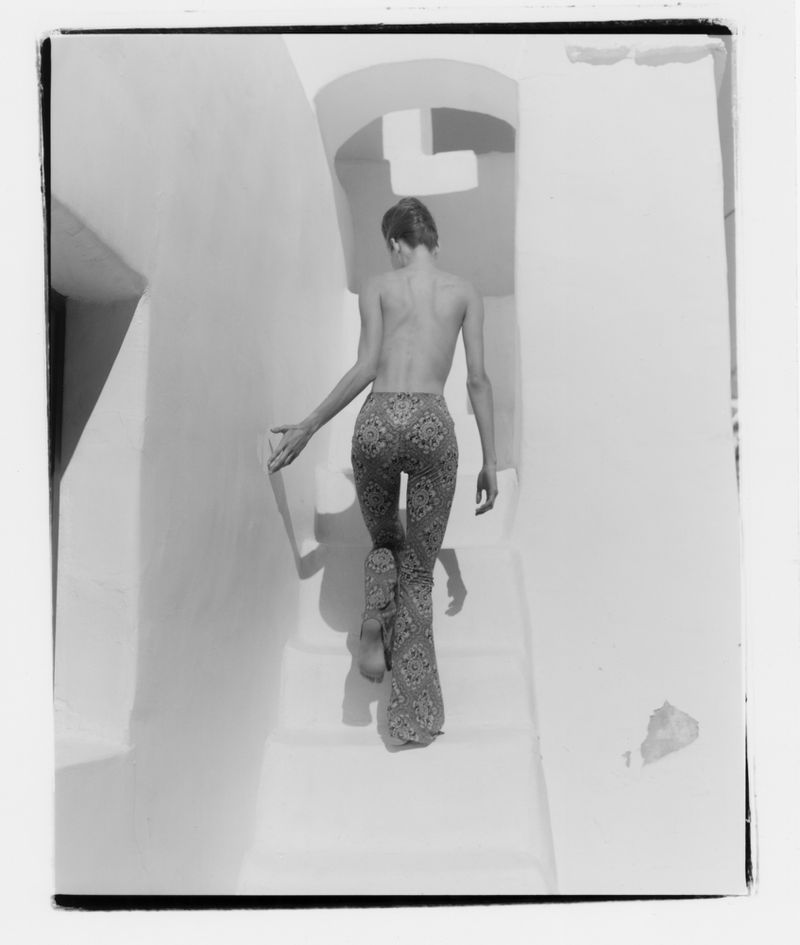 Back view of a woman in patterned pants climbing up a white staircase with arches and white walls in a black and white photograph.