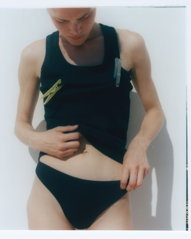 A woman in a black tank top and bikini bottom stands against a white wall, holding the bikini bottom, with clothing pins on the tank top.