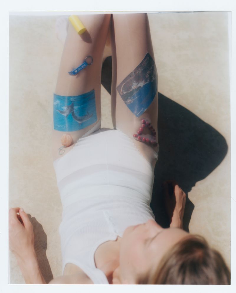 Blonde woman lying on beige fabric, legs up, wearing a white shirt with stickers and toys on her legs in an outdoor setting.